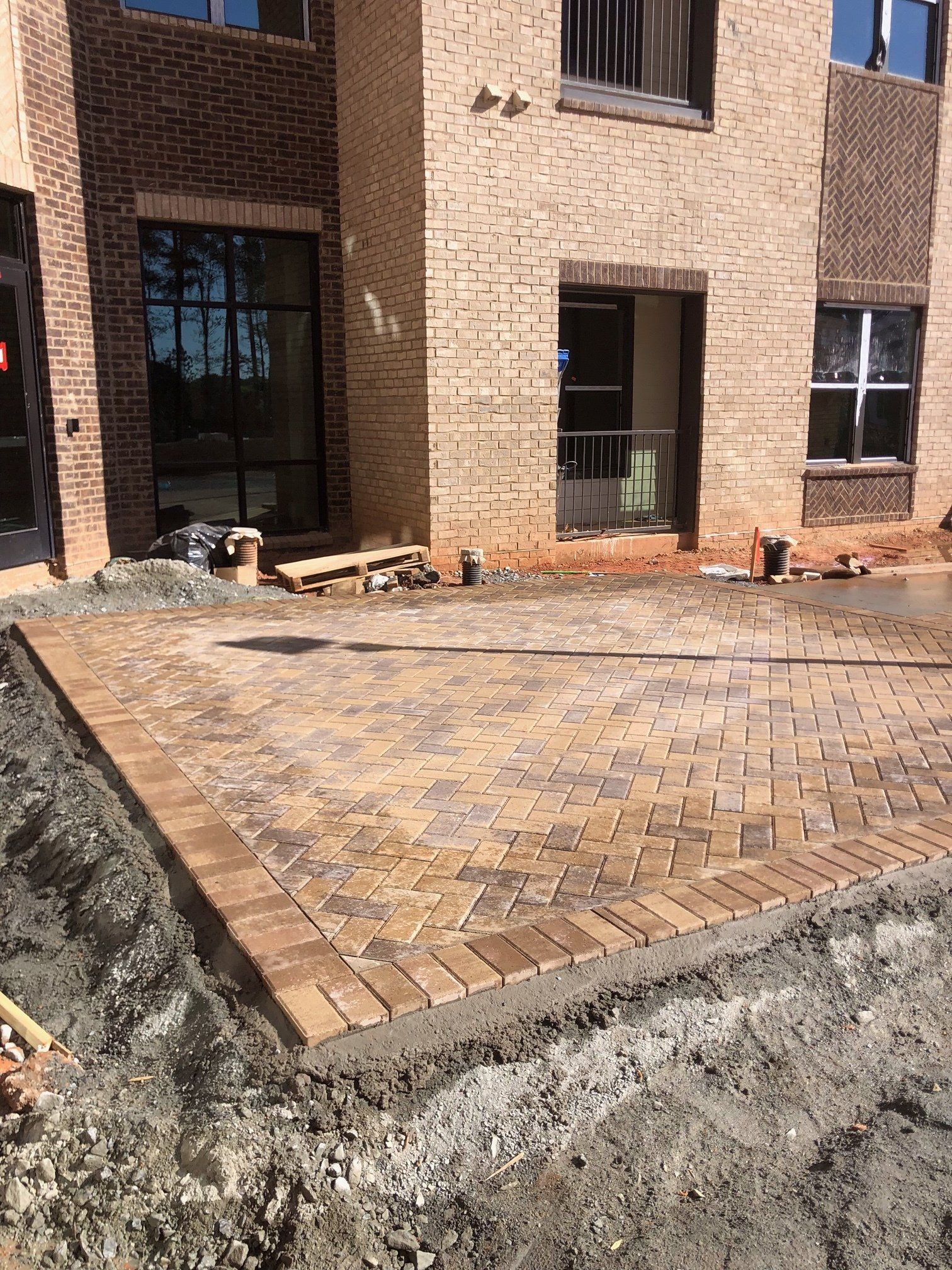 Brick patio under construction beside a building, with unfinished concrete edges and stacked materials nearby