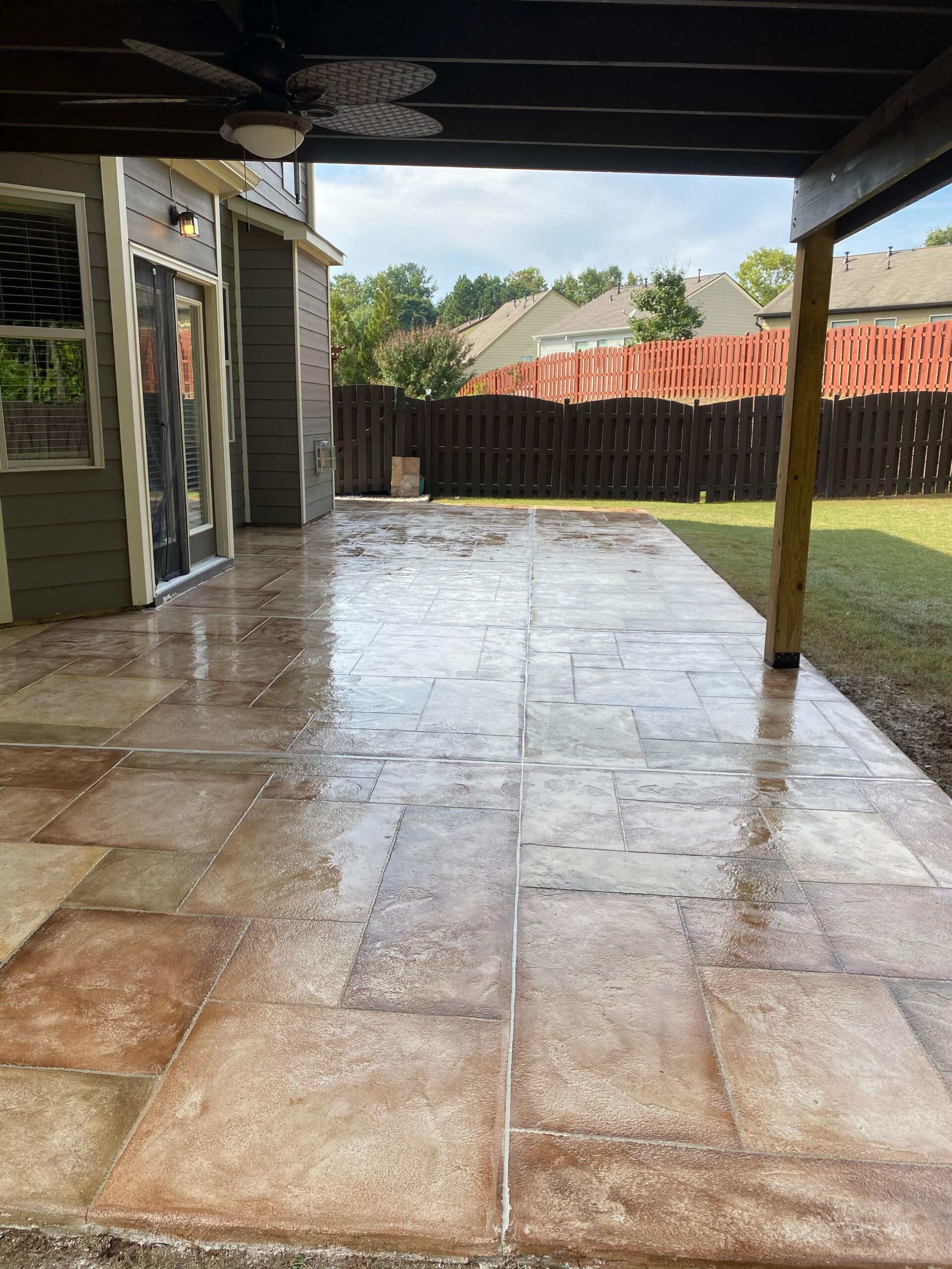 Covered patio with wet stone floor, wooden posts, and backyard fence after rain