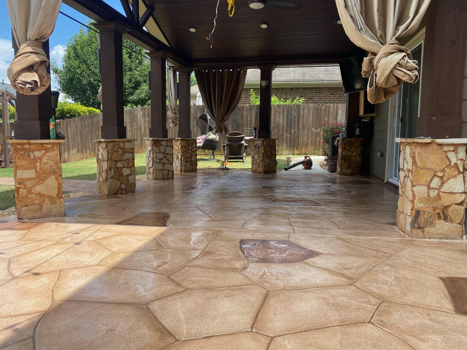 Covered outdoor patio with stone columns, tiled floor, and draped curtains overlooking a backyard fence.