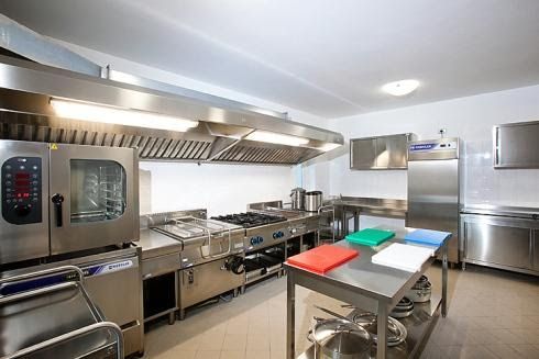 Cucina professionale in acciaio inox con forni, piani cottura, tavolo di preparazione e cappa aspirante.