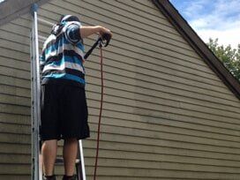 Person on a ladder spraying siding of a house with a red hose.