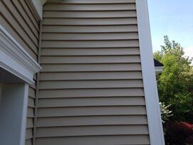 Tan siding on a house corner with white trim. Trees and blue sky visible in the background.