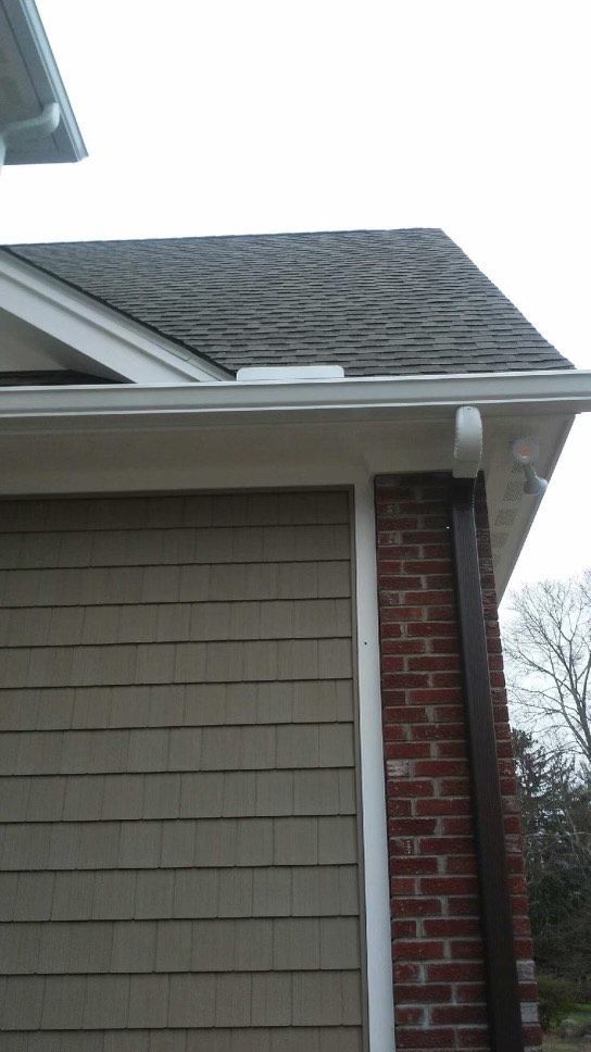 Corner of a house with beige siding, brick, and a dark roof with gutters and a downspout.