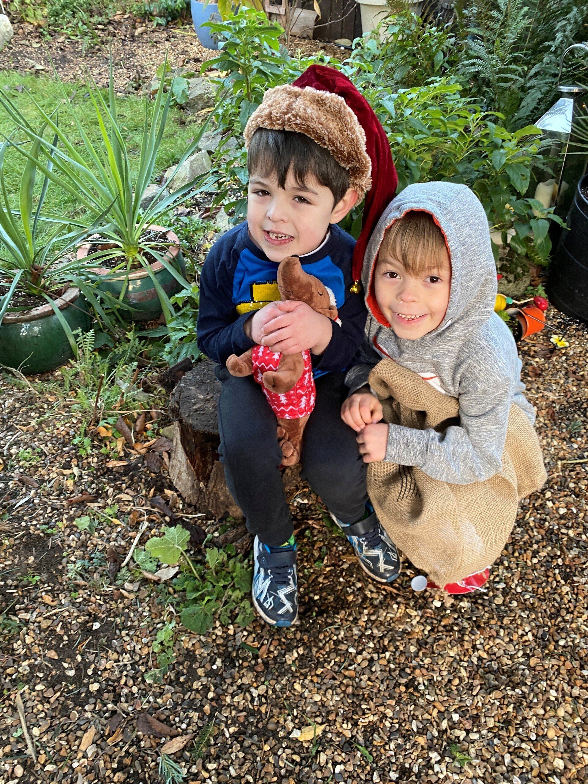 Two children in a garden, one wearing a Santa hat, both smiling and holding stuffed animals.