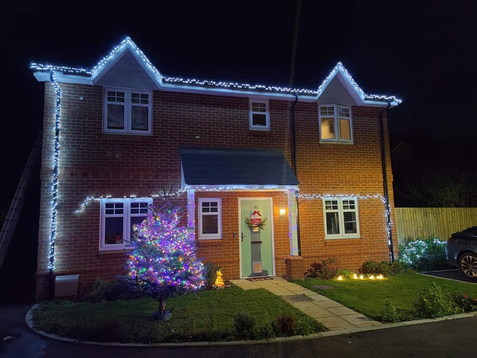 Two-story brick house with Christmas lights outlining roof and windows. Lit Christmas tree on the lawn.