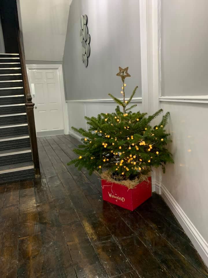 Christmas tree with fairy lights in a red box on a wooden floor near a staircase.