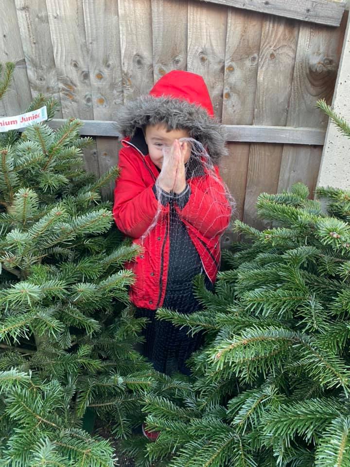 Child in red coat among Christmas trees, holding hands near face, smiling. Wooden fence background.