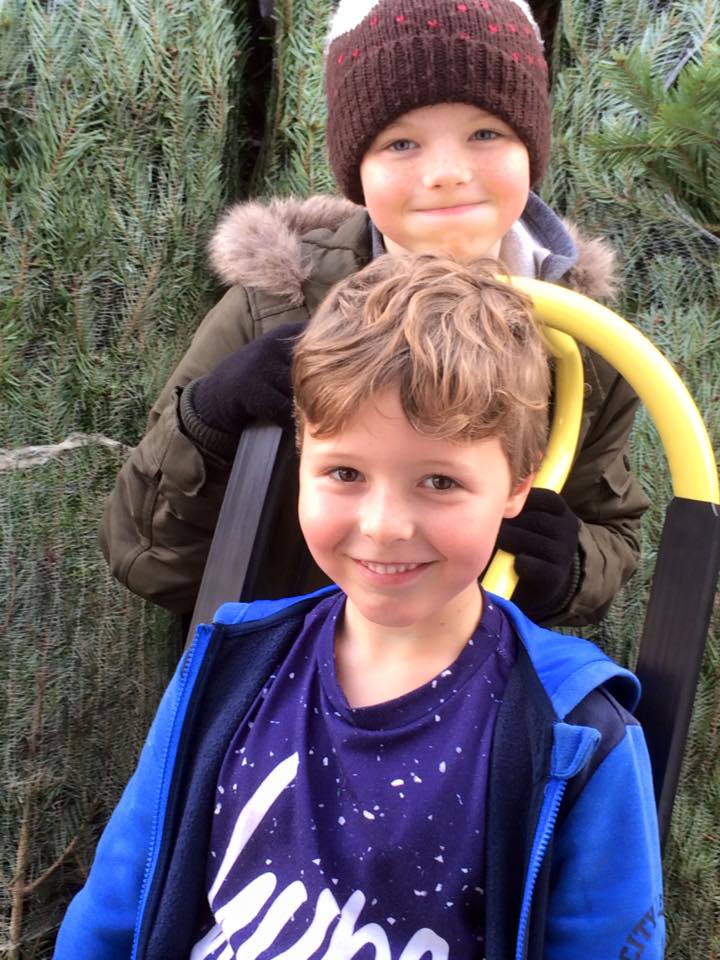 Two boys smiling near a stack of evergreen trees. One wears a blue jacket, the other a winter hat and olive jacket.