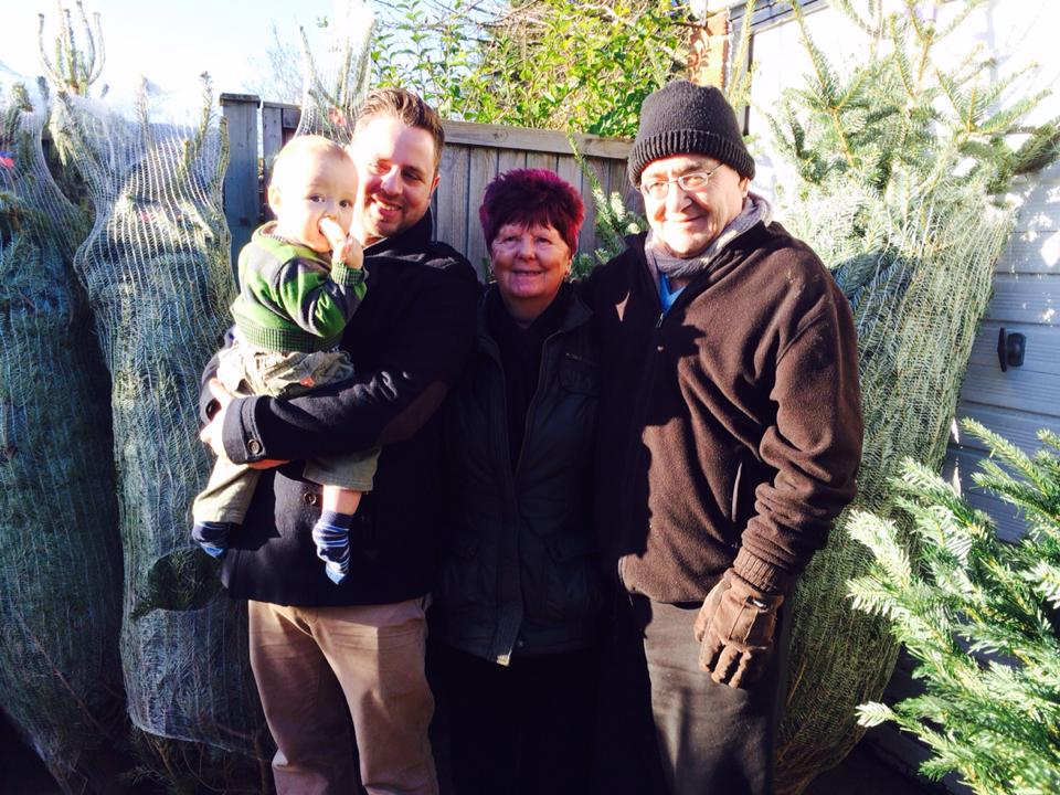 Family of four poses in front of wrapped Christmas trees; child held by man, other adults smile.