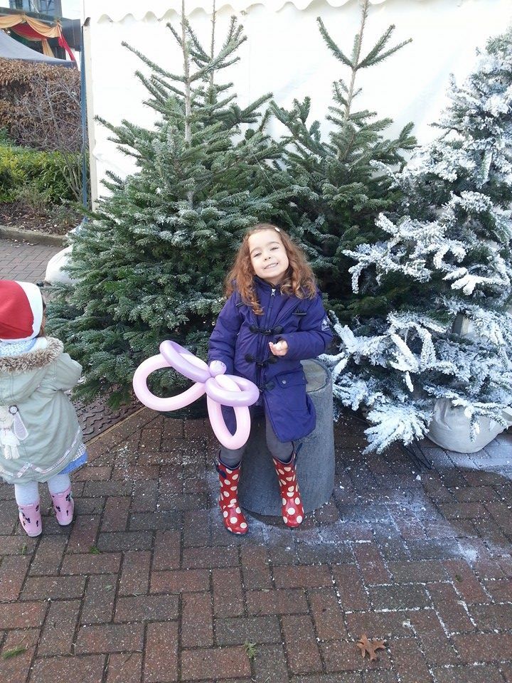 Girl in a purple coat and red boots holds a pink balloon near Christmas trees.