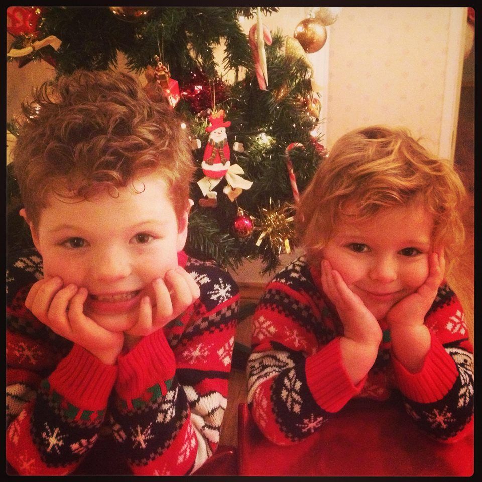 Two children in matching holiday sweaters pose in front of a decorated Christmas tree, smiling with hands on their chins.