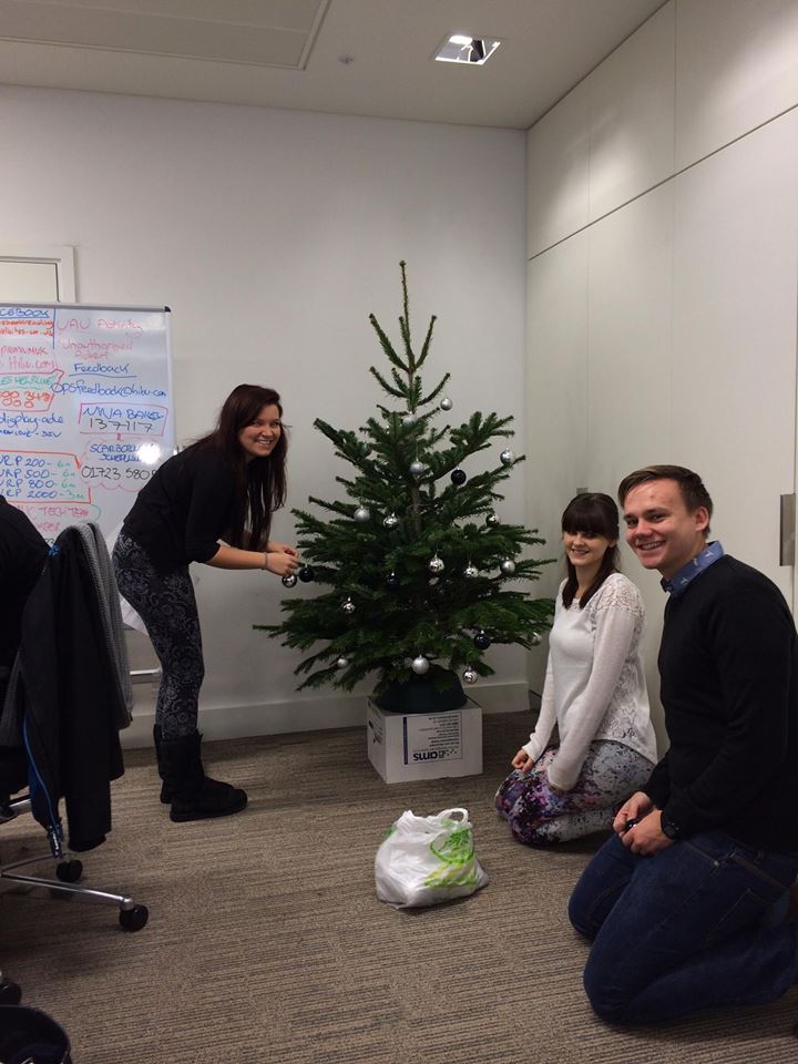 Three people decorate a Christmas tree in an office. One woman holds an ornament, two kneel on the floor.