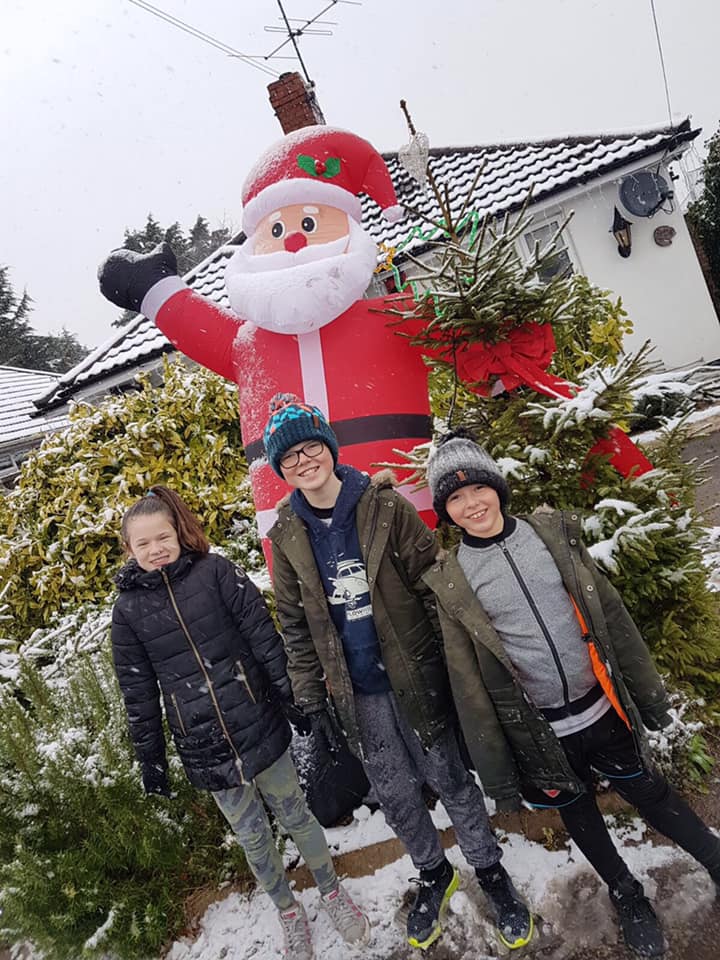Three children pose in front of a giant inflatable Santa in a snowy yard.