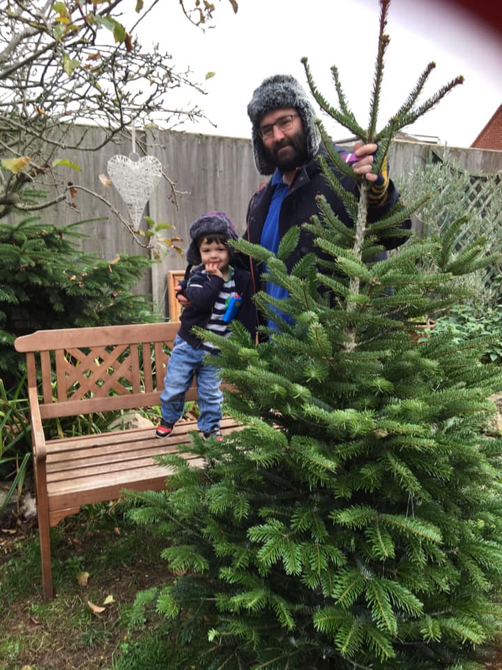 Man and child in winter hats pose with a Christmas tree in a garden.