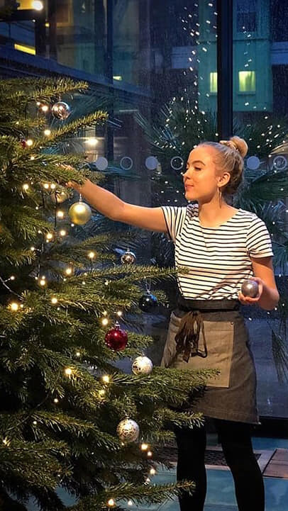 Woman decorating a Christmas tree with ornaments, wearing a striped shirt and apron in a building.