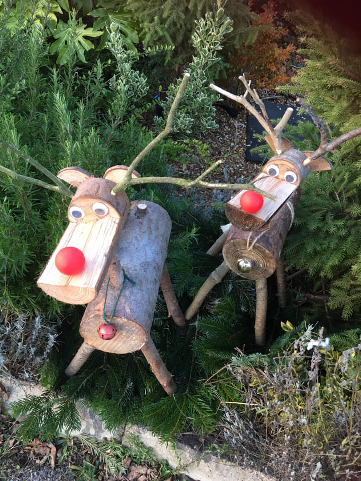 Two wooden reindeer decorations with twig antlers and red noses, in a garden setting.
