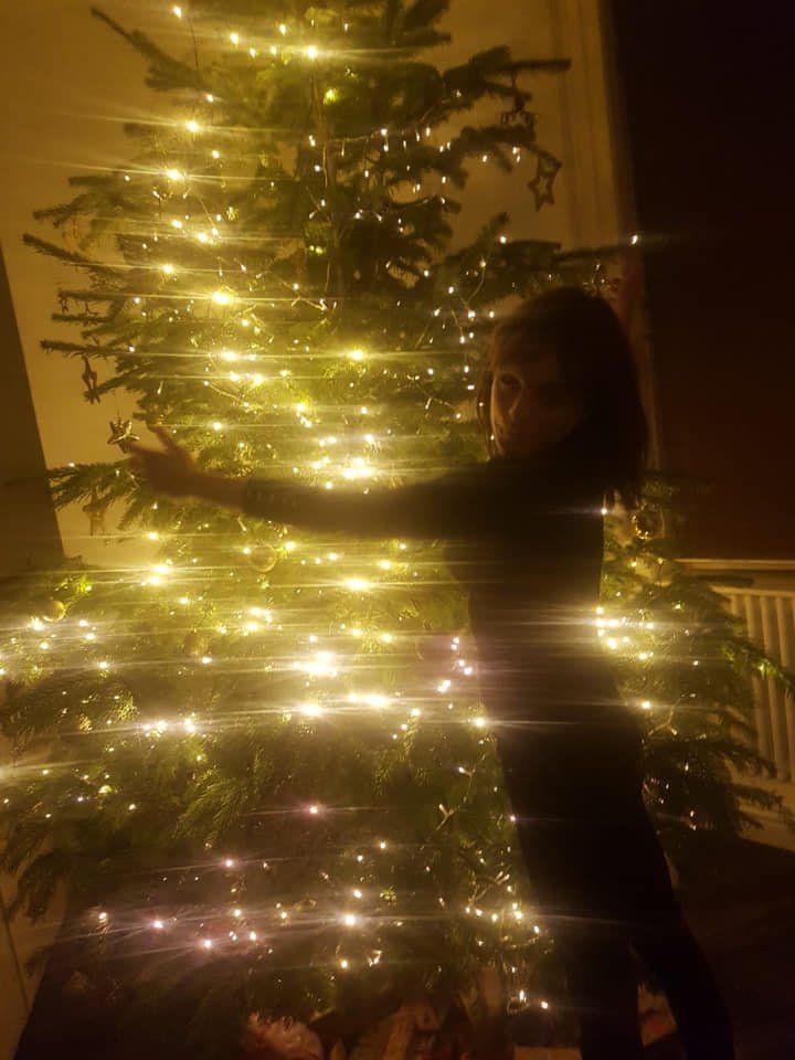 Child stands near a brightly lit Christmas tree with outstretched arms, indoors at night.
