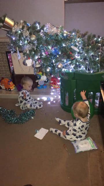 Two toddlers in pajamas play near a Christmas tree, decorations scattered.