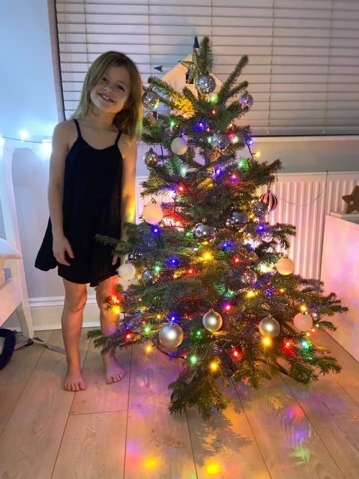 A girl smiles next to a decorated Christmas tree with colorful lights indoors.