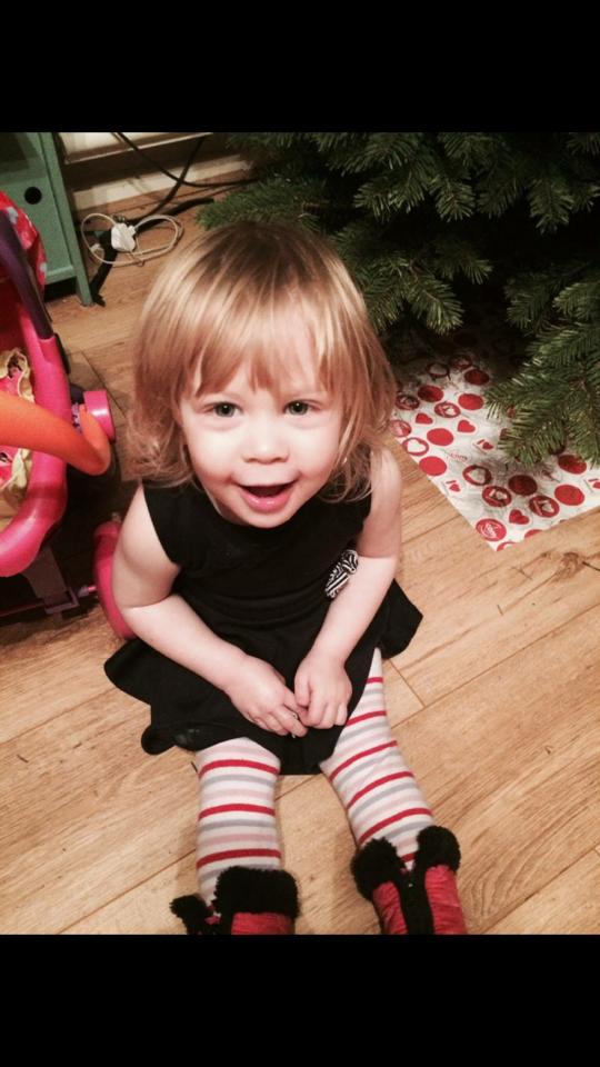 Young child with blonde hair, smiling, wearing a black dress and striped socks, seated near a Christmas tree.