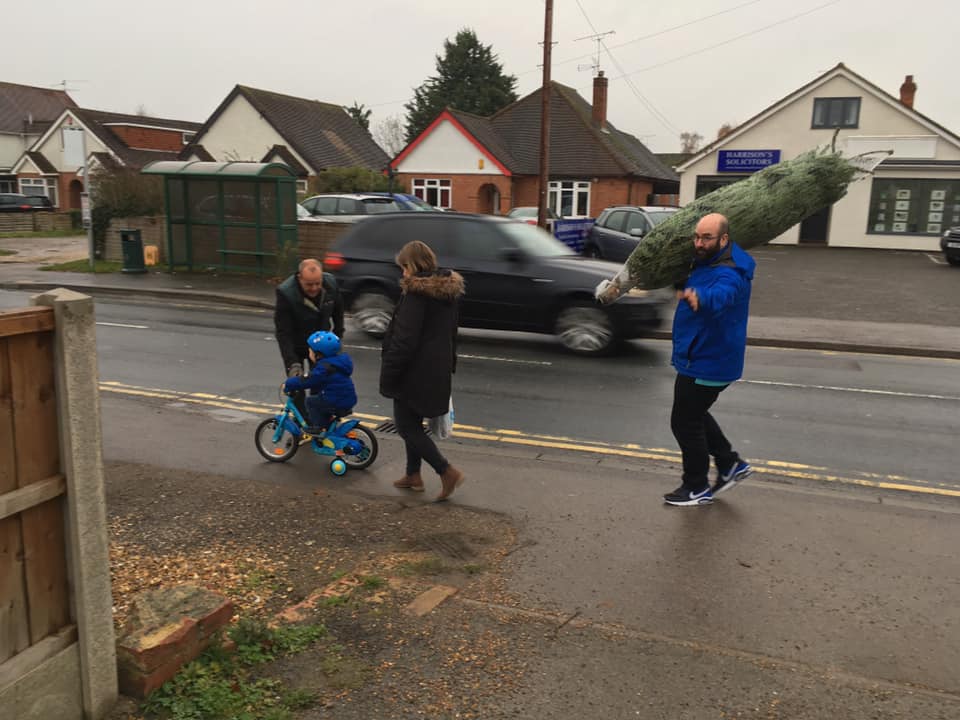 Man carrying Christmas tree, family on sidewalk near road. Child on bike, others walking. Overcast day.