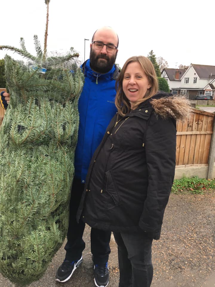 Couple holding a wrapped Christmas tree outdoors, near a fence, looking at the camera.