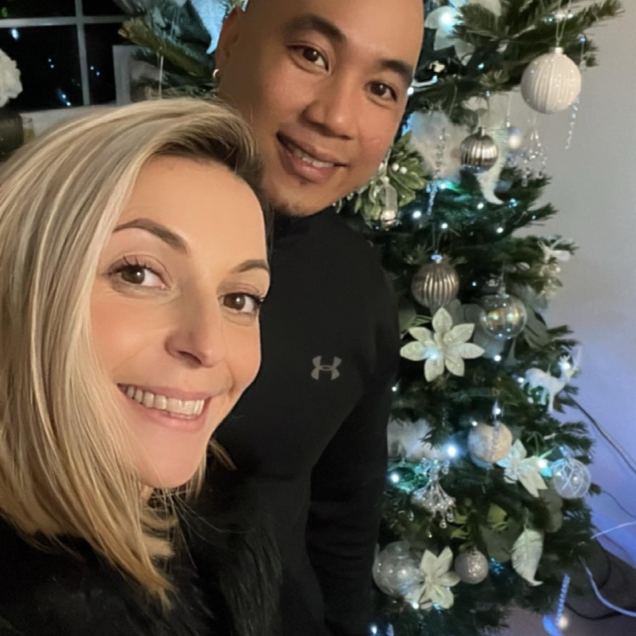 Couple smiling in front of a decorated Christmas tree with lights and ornaments.