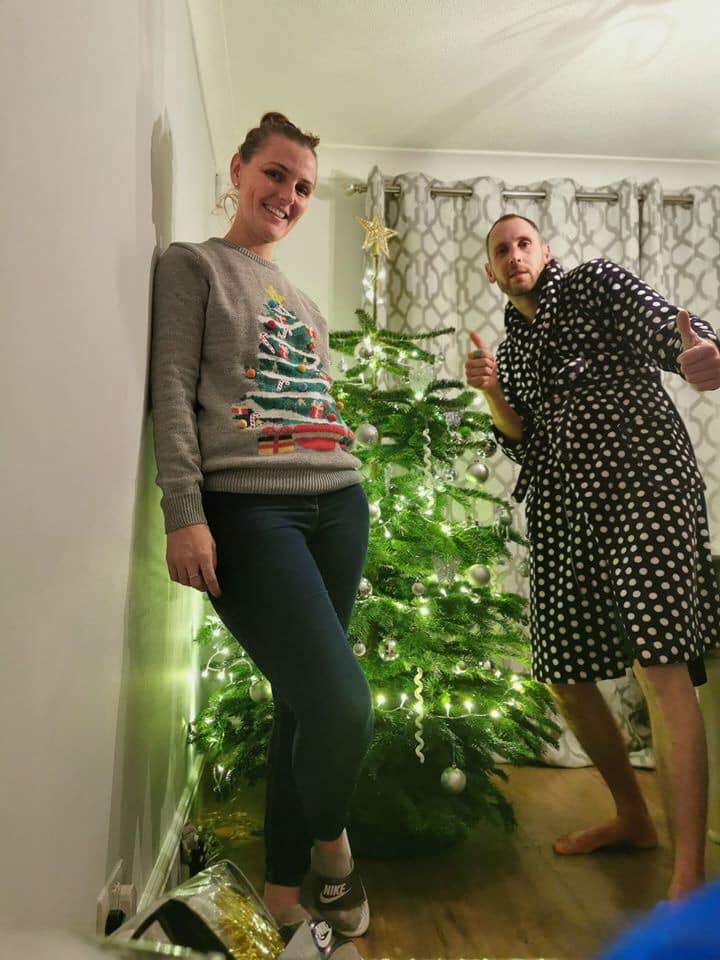 Woman leans against wall, wearing Christmas sweater; man in robe gives thumbs up next to decorated tree.
