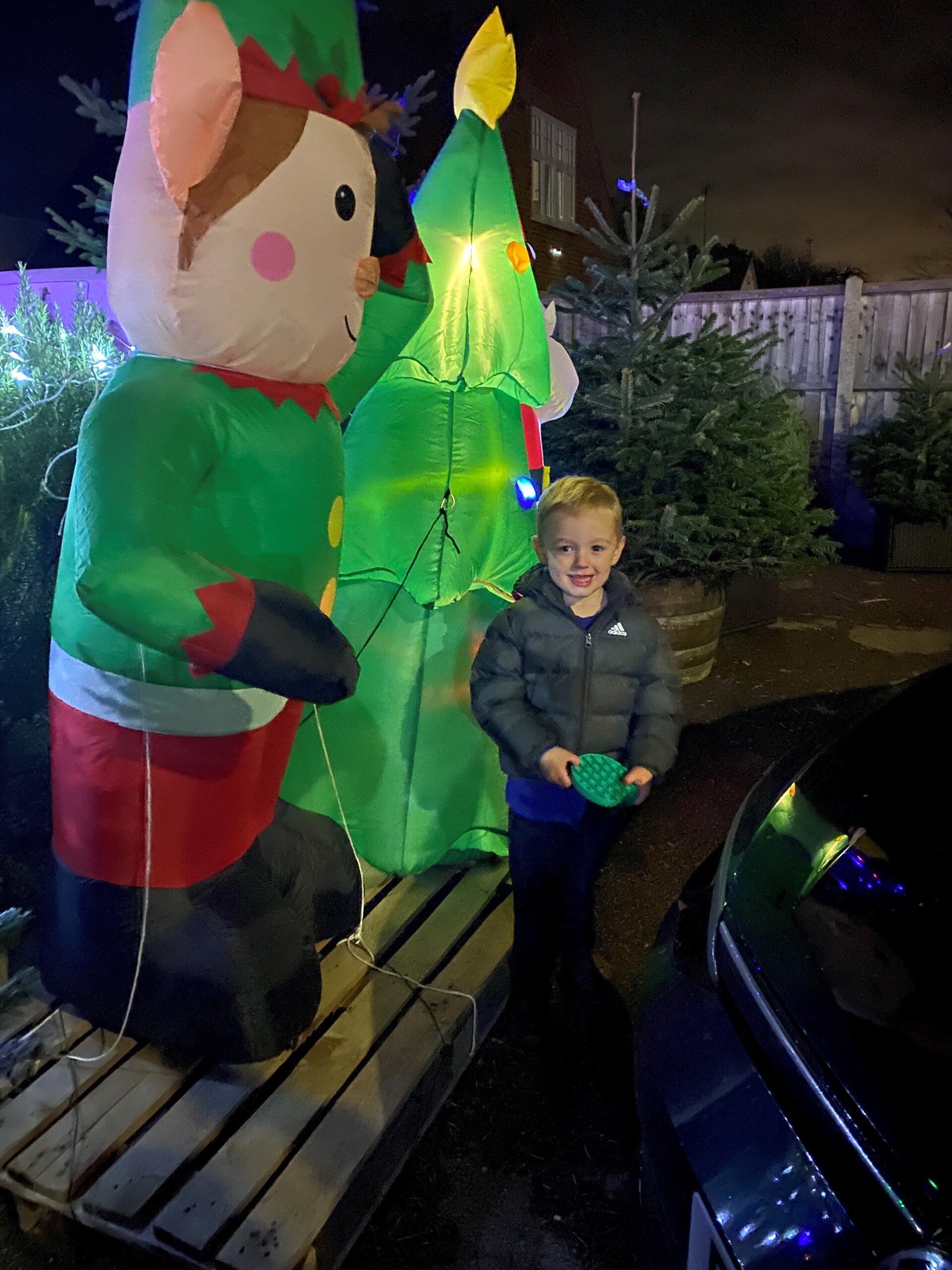 Boy stands with an inflatable elf and tree; nighttime outdoor setting, lights visible.