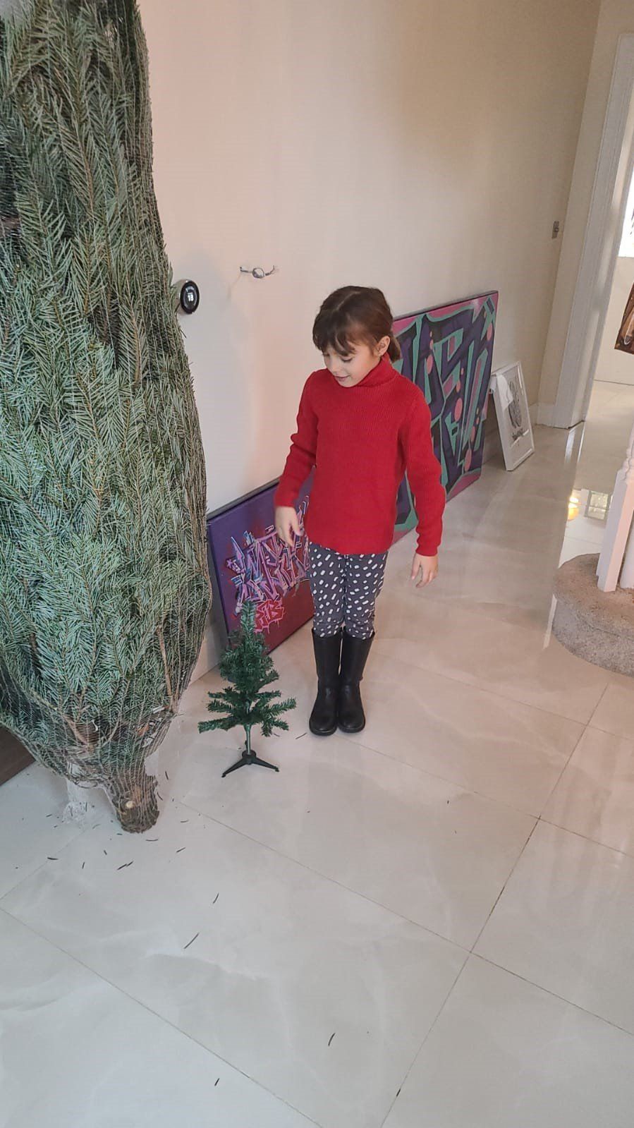 Child in red sweater and dotted pants with a small tree near a large, bare Christmas tree.