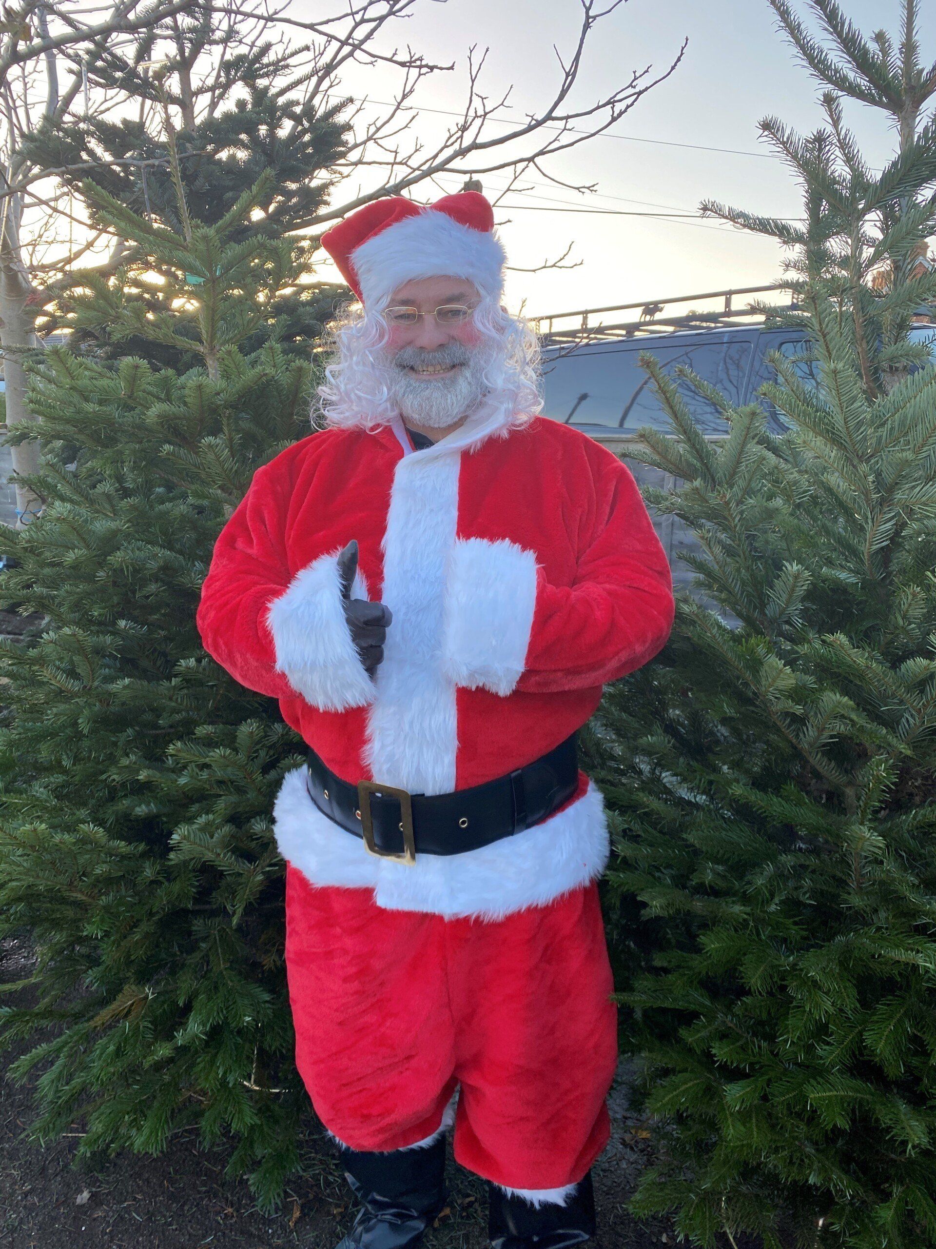 Santa Claus standing outdoors, smiling, with Christmas trees in the background.