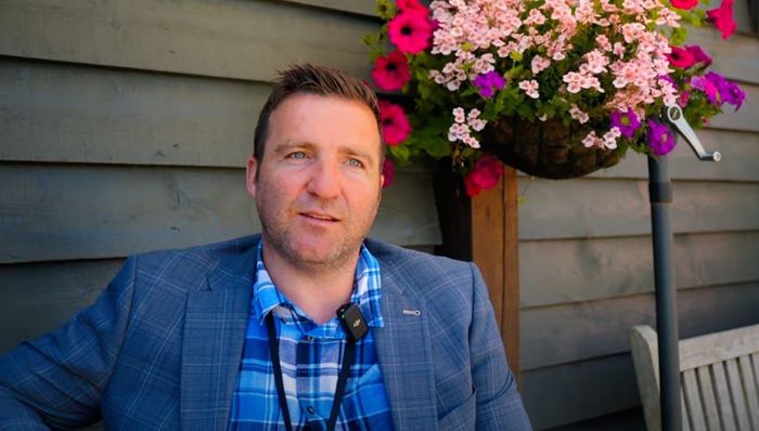 Man wearing blue blazer and checked shirt, sitting outside, talking. Flowers in hanging basket.