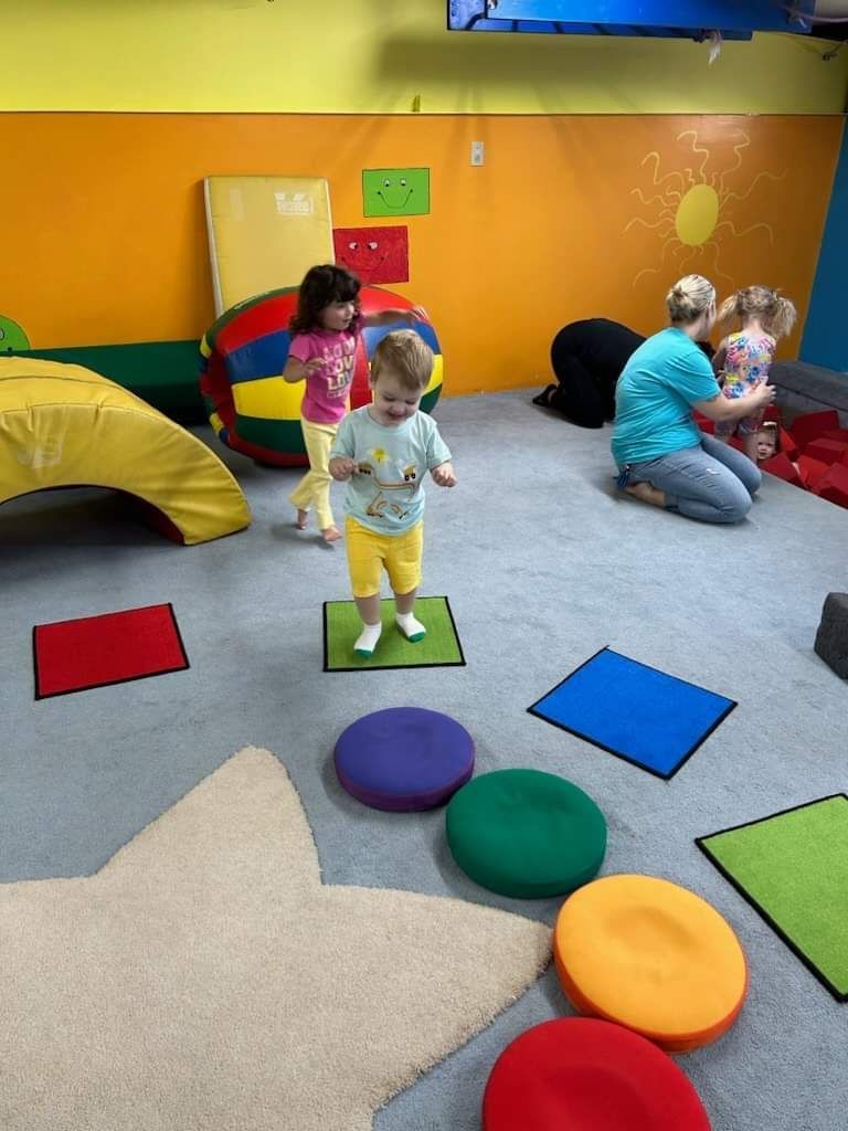 Children playing in a colorful play area; one child stands on a green mat, adults supervise.
