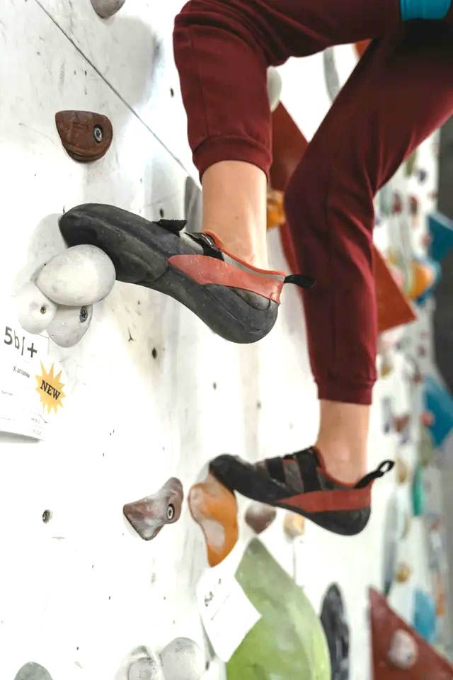 Person rock climbing, feet on holds. Red and black climbing shoes, red pants, white wall.