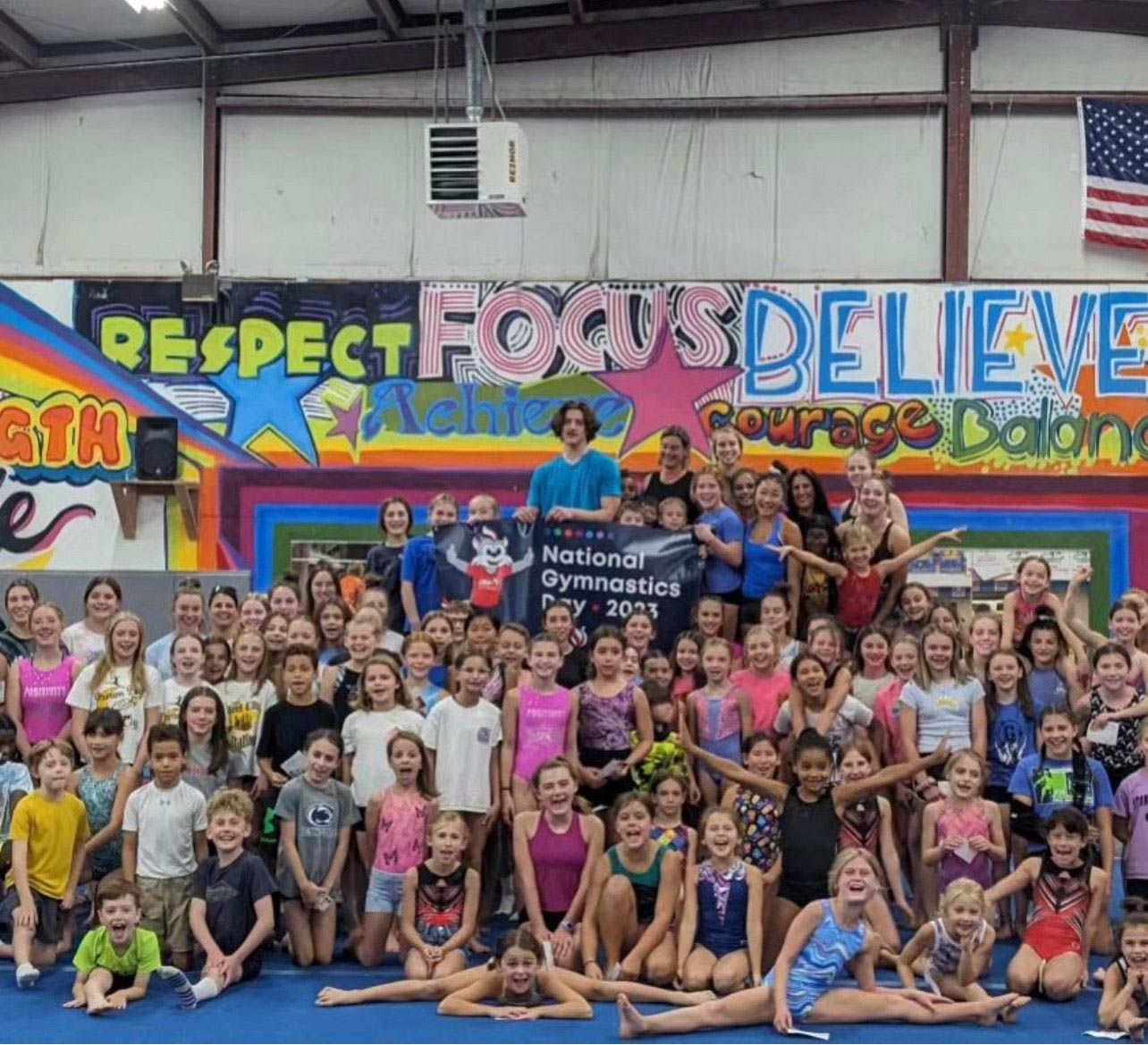 Large group of gymnasts smiling together; in a gym for National Gymnastics Day, blue floor.