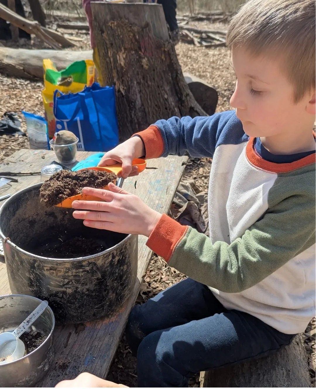 Boy using a trowel to scoop soil into a metal pot outdoors.