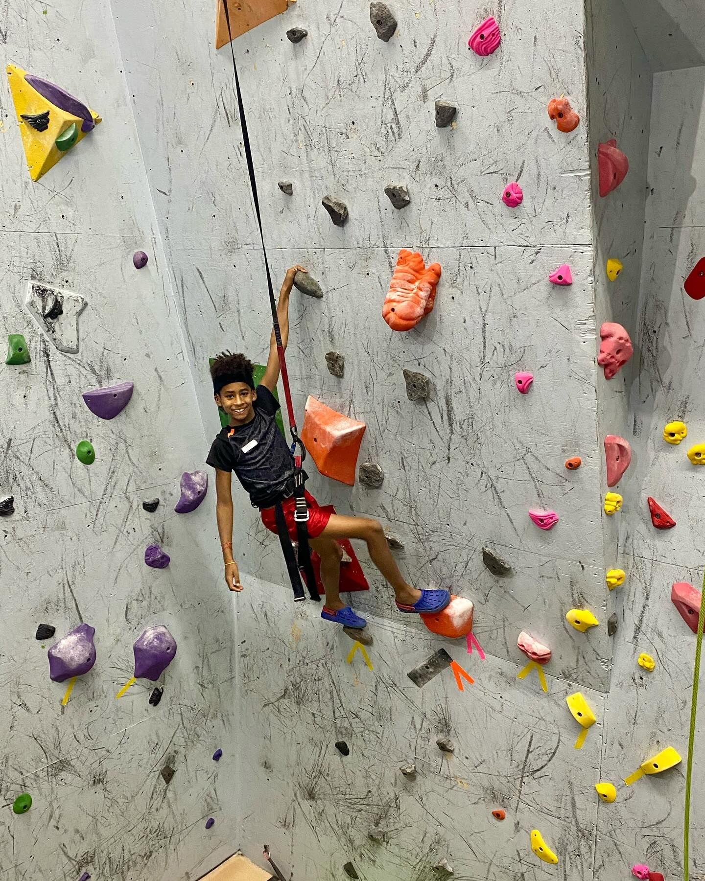 Woman climbing an indoor rock wall, legs dangling, arms stretched. Colorful handholds on a textured wall.
