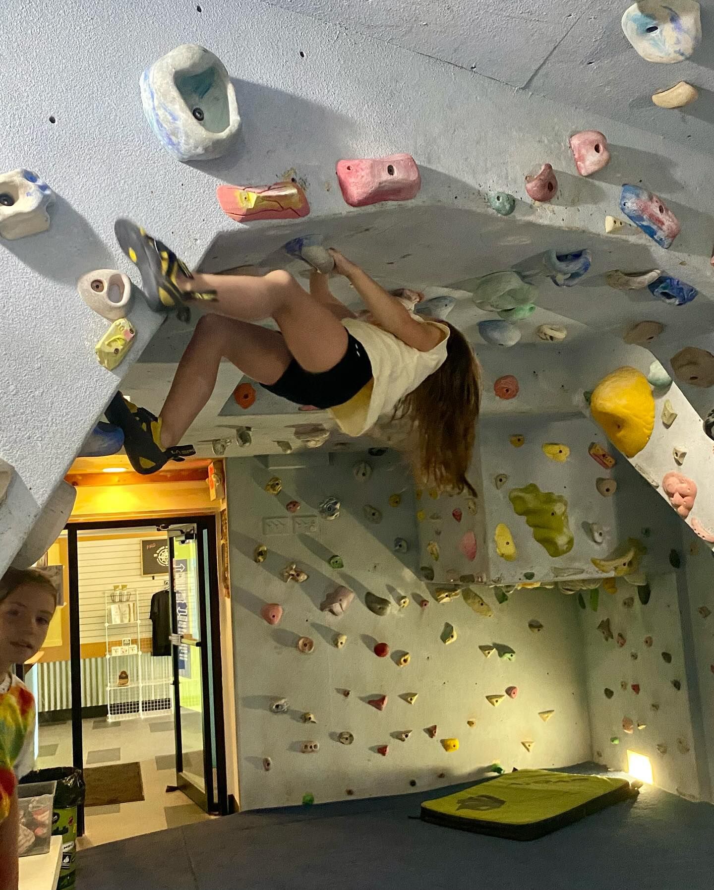 Woman climbing an indoor rock wall, legs dangling, arms stretched. Colorful handholds on a textured wall.