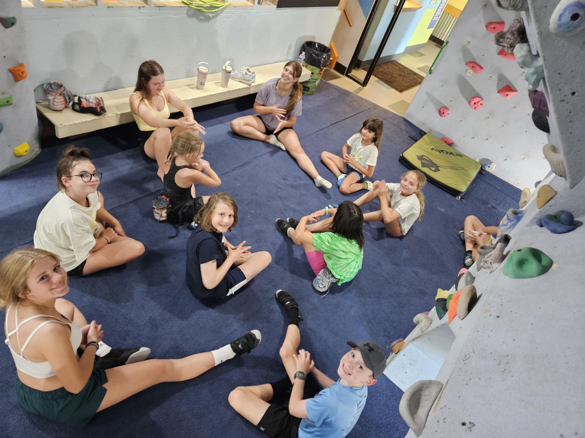 Group of diverse kids sitting on blue mat at climbing gym, smiling and looking toward the camera.