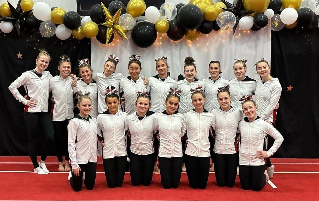 Cheerleading team in white and black uniforms posing on a red mat, with balloons behind them.