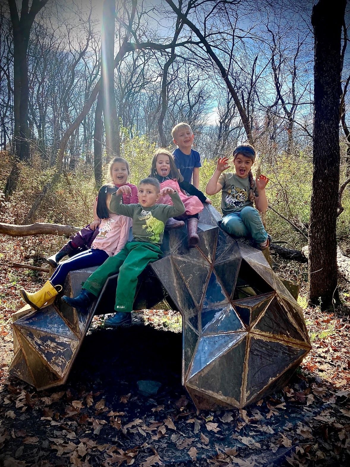Six children, smiling, sitting on a metal sculpture in a wooded area.
