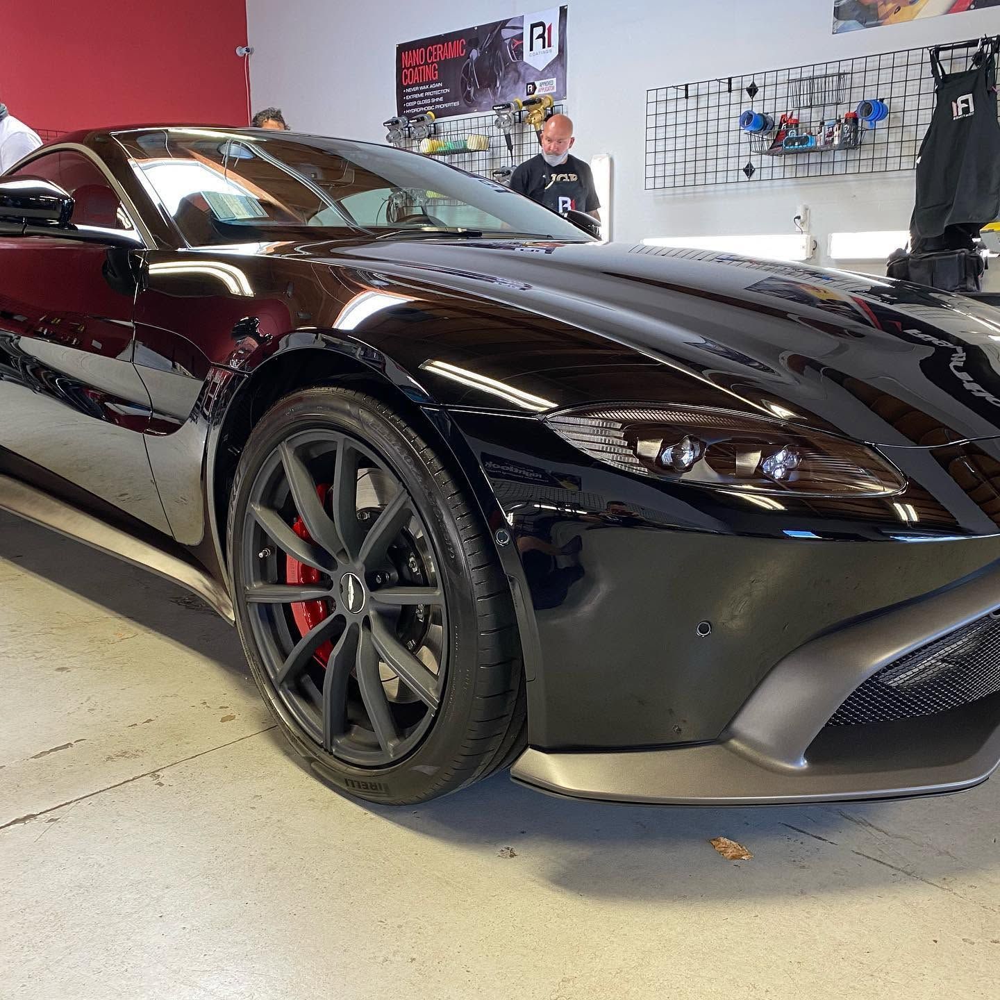 Glossy black sports car on display indoors, with red brake calipers and people in the background.