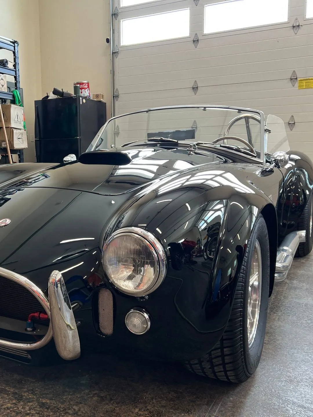 Black AC Cobra roadster parked indoors. Front view. Chrome accents, silver wheels, and a windshield.