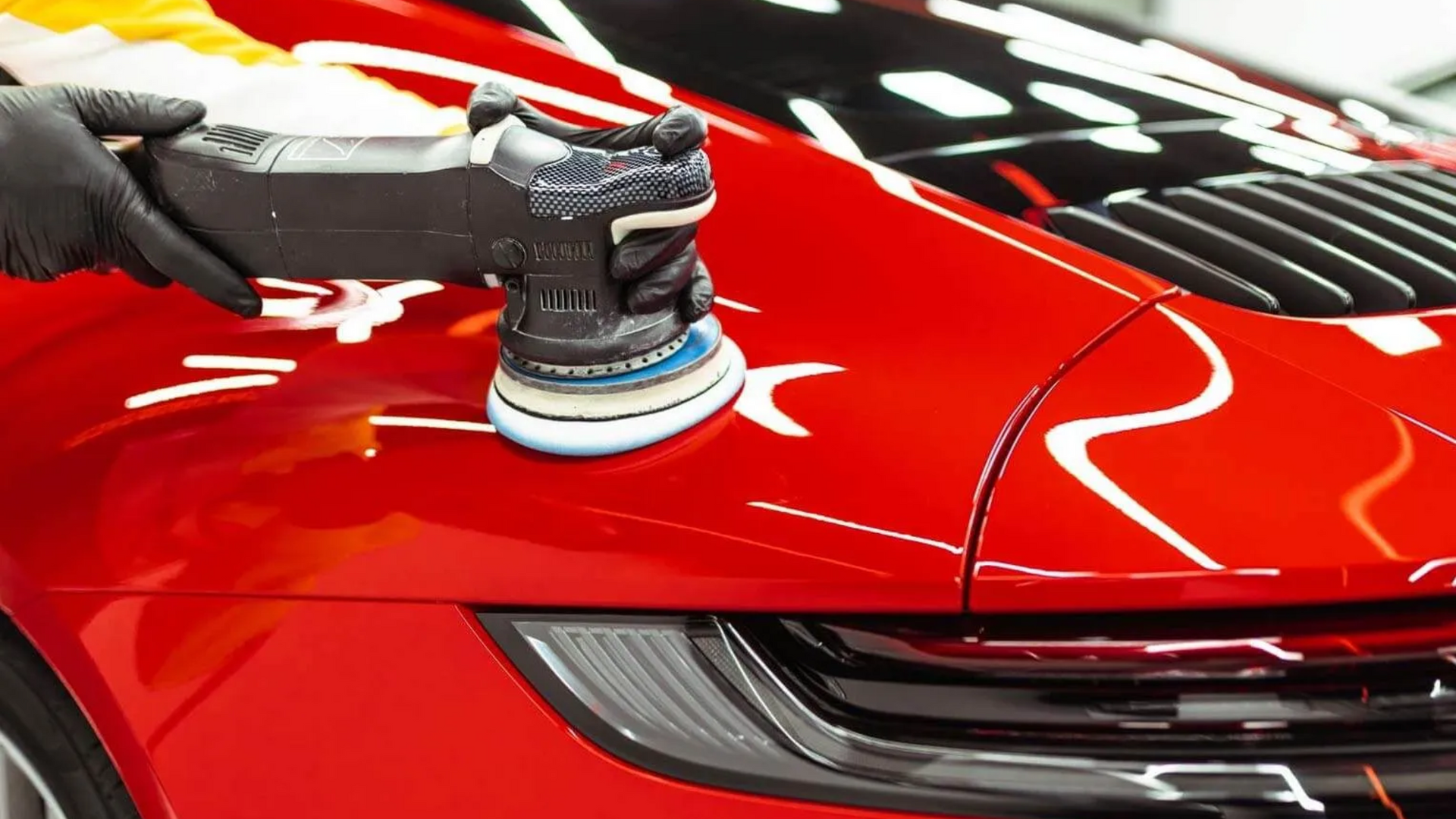 Black-gloved hand using a polisher on a red car's body. The car is outdoors, with bright reflections on the surface.