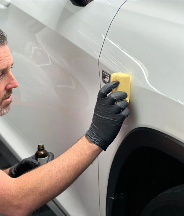 Man applying liquid to a white car with a yellow applicator; black gloves.