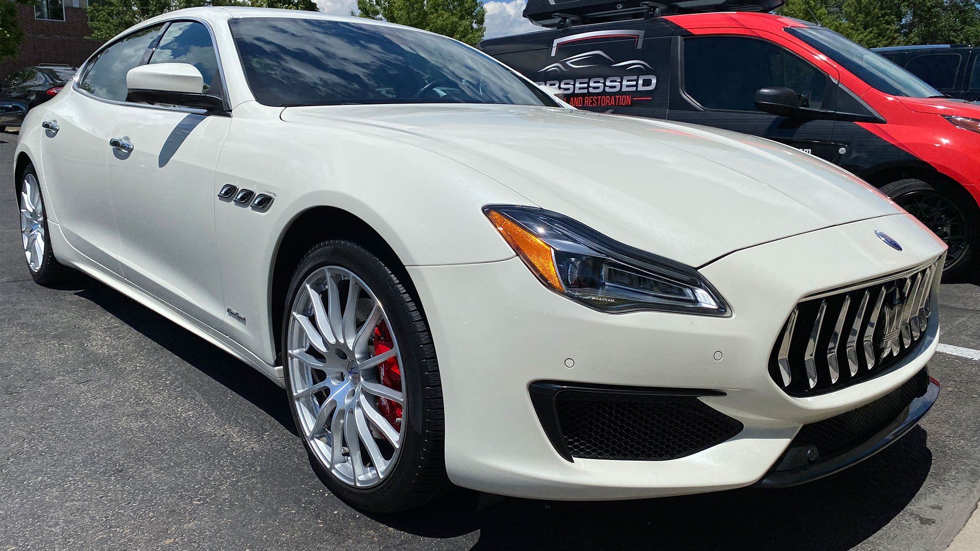 A white maserati is parked next to a red car in a parking lot.