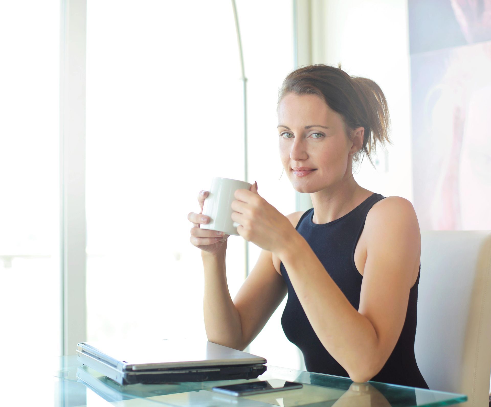 Woman holding a white mug at a glass table with a laptop, in a bright room