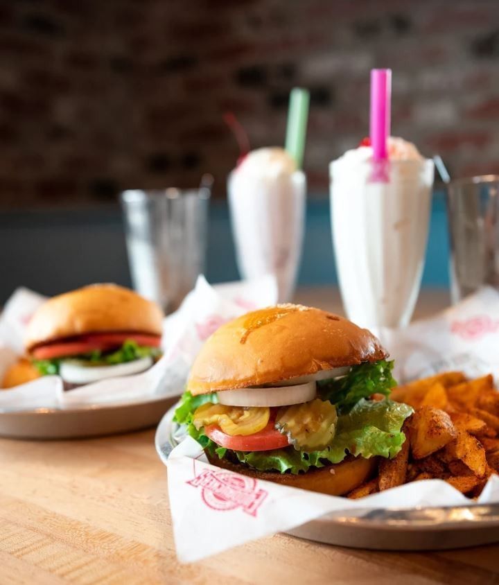 A hamburger is on a plate with french fries and milkshakes in the background
