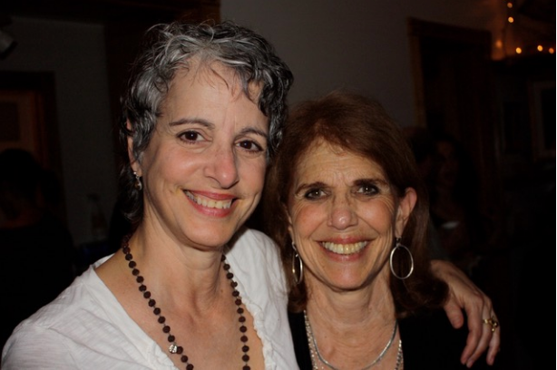 Two smiling women, one with short gray hair, the other with long auburn hair, arms around each other, indoors.