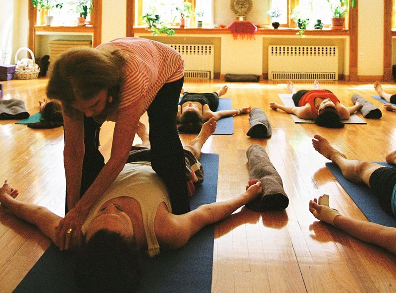 Yoga instructor assisting a student in a pose; others relax on mats in a bright studio.